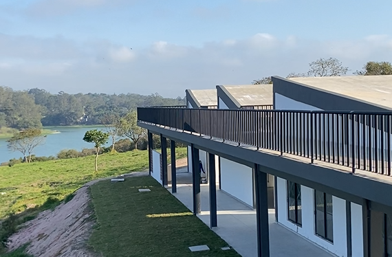A imagem mostra uma construção moderna de linhas retas, pintada em branco com detalhes cinza-escuros, incluindo colunas e corrimão metálico no andar superior. O prédio tem grandes janelas de vidro e está localizado em uma área verde, com gramado em primeiro plano e um lago de água azul-esverdeada ao fundo, rodeado por árvores e vegetação. O céu aparece parcialmente nublado, em tons de azul claro com nuvens brancas, compondo uma paisagem natural harmoniosa ao redor da edificação.