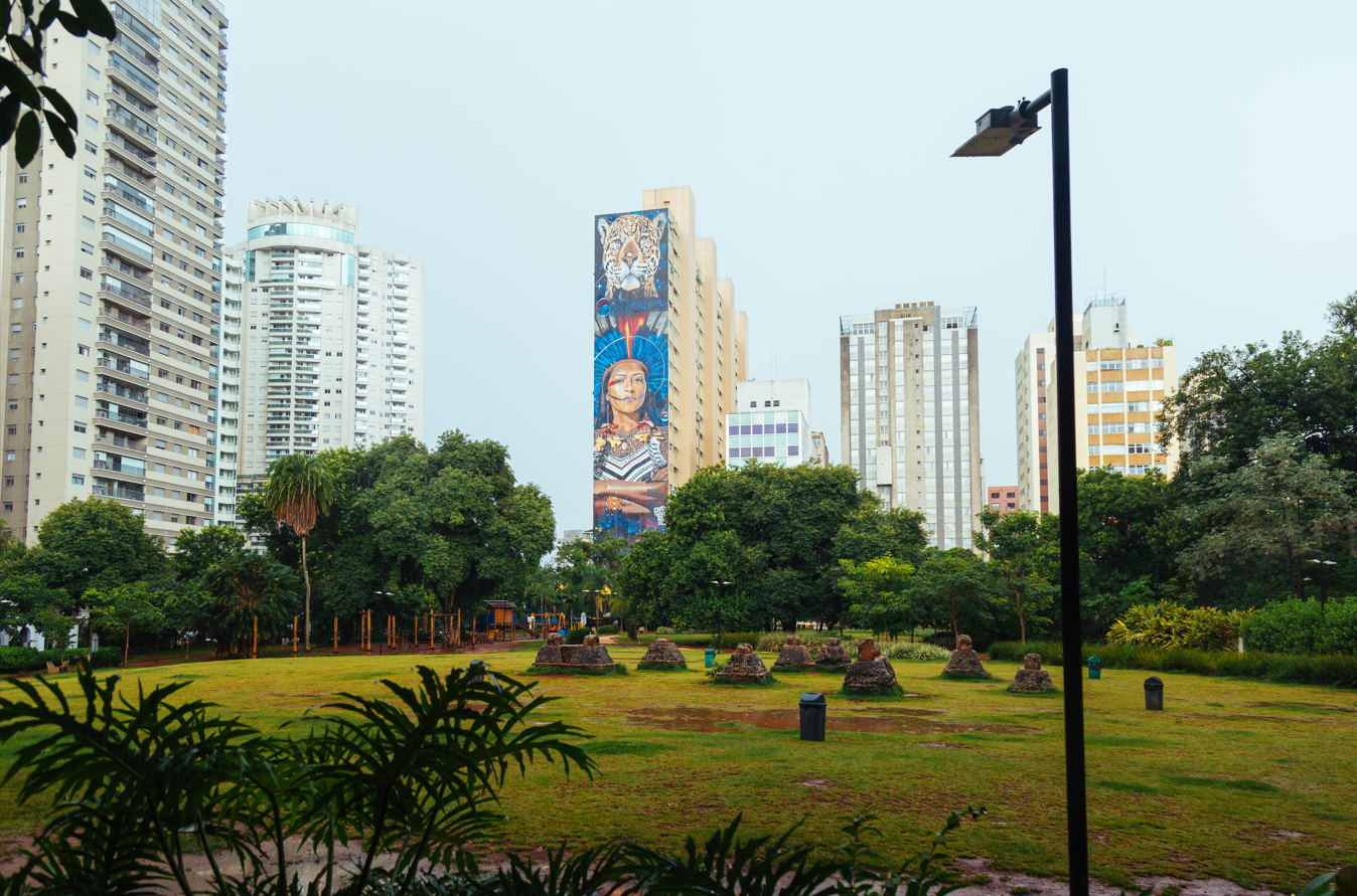 A imagem captura uma cena urbana que contrasta um espaço verde com um horizonte de edifícios altos. Em primeiro plano, domina um gramado verde, levemente molhado ou com poças d'água, sugerindo chuva recente ou orvalho. Sobre este gramado, estão dispostas fileiras de estruturas baixas e arredondadas, que parecem ser pequenos monumentos ou formações rochosas, algumas cobertas por musgo ou terra. Folhagens exuberantes emolduram a parte inferior da cena, criando uma profundidade natural em tons de verde escuro e verde claro. No centro da composição, destaca-se um poste de luz preto, com um design moderno.  Ao fundo, o cenário é dominado por uma paisagem arquitetônica densa sob um céu claro e esbranquiçado. Vários arranha-céus de tons predominantemente bege, cinza e branco se elevam, mas o ponto focal da paisagem urbana é um edifício central que ostenta um mural impressionante. Este mural vibrante retrata uma mulher indígena e uma onça-pintada, utilizando cores ricas como azul profundo, laranja, vermelho e tons terrosos. A vegetação densa e variada, em tons de verde, forma uma barreira natural entre o parque e a estrutura urbana.
