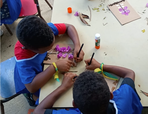 Imagem vista de cima mostra duas crianças sentadas à mesa, participando de uma atividade manual. Elas usam camisetas azuis e pulseiras verdes e amarelas nos pulsos. Sobre a mesa clara, as crianças colam pétalas de flores roxas, folhas secas e pequenos galhos em pedaços de papel marrom, usando cola branca e cola em bastão. Uma das crianças segura um lápis enquanto ajusta os elementos naturais no papel. Ao redor, há restos de folhas, pétalas espalhadas e materiais de arte. O ambiente parece ser um espaço escolar ou educativo ao ar livre, com clima de concentração e criatividade.