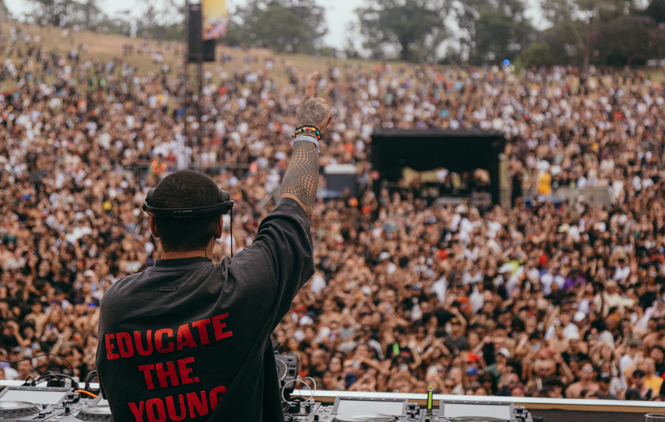 Imagem colorida, em plano aberto, mostrando um grande show ao ar livre durante o dia. Em primeiro plano, de costas para a câmera, há um DJ usando fones de ouvido e camiseta preta com a frase “EDUCATE THE YOUNG” escrita em vermelho nas costas. Ele levanta um dos braços, com o dedo indicador apontado para o alto, em gesto de animação e comando da plateia. O braço tem tatuagens visíveis e pulseiras coloridas no pulso. À frente dele, sobre a mesa, estão equipamentos de DJ, como controladores e cabos.  Ao fundo, vê-se uma multidão muito grande, composta por milhares de pessoas, espalhadas por uma área ampla e levemente inclinada, parecida com um gramado ou parque. O público está bem próximo uns dos outros, muitos com os braços levantados, acompanhando o show. Mais atrás, há um palco coberto e árvores espalhadas, indicando um evento em área aberta. A atmosfera é vibrante, festiva e cheia de energia coletiva.