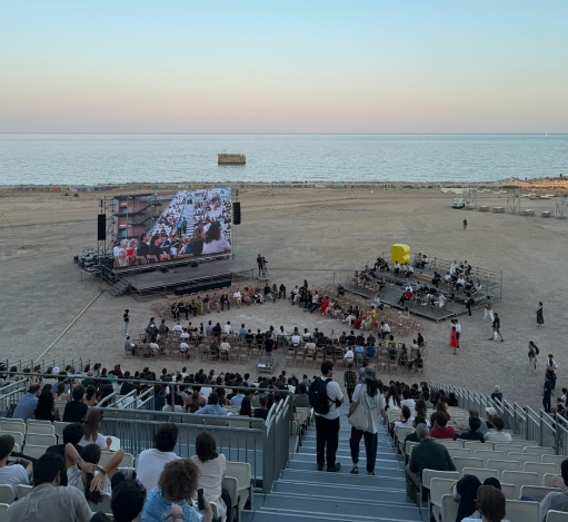 **Para cego ver:** Fotografia registrada do alto de uma arquibancada montada na praia, durante o entardecer, em Barcelona, na Espanha. A imagem mostra a cerimônia do **Congresso Mundial de Arquitetos UIA 2026**, realizada à beira-mar. No centro da arena, há um palco com um grande telão exibindo imagens do evento, cercado por participantes sentados em formato circular. Nas arquibancadas, dezenas de pessoas acompanham a programação. Ao fundo, o mar calmo se encontra com o horizonte sob um céu em tons suaves de azul e rosa, criando um cenário amplo e iluminado pela luz natural do fim da tarde.