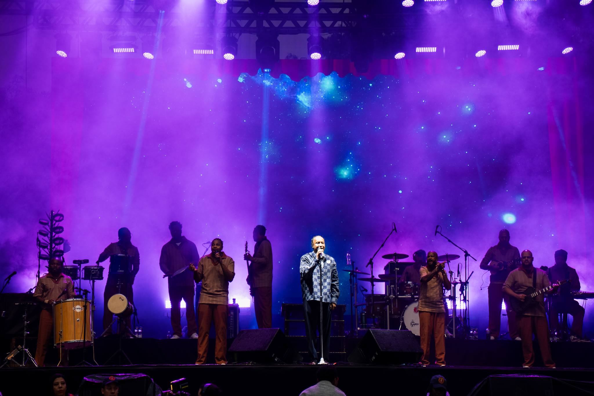 Para cego ver: palco de show ao ar livre durante a Virada Cultural, iluminado por luzes em tons de roxo e azul. Ao centro, um cantor se apresenta acompanhado por diversos músicos distribuídos pelo palco, com instrumentos de percussão, bateria e guitarras. Ao fundo, um grande painel de LED exibe imagens que lembram o céu estrelado e o espaço sideral. A fumaça das luzes cria um efeito vibrante e envolvente, enquanto a plateia acompanha a apresentação em frente ao palco.