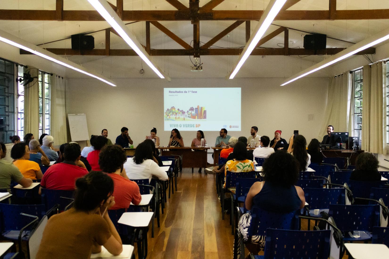 Imagem de uma sala de reunião ou auditório. O espaço é amplo, com piso de madeira e teto alto, onde vigas de madeira ficam aparentes. Longas luminárias brancas estão suspensas no teto, iluminando o ambiente.  Em primeiro plano, várias pessoas estão sentadas em cadeiras organizadas em fileiras, voltadas para a frente. Elas assistem a uma mesa de debate composta por cerca de oito pessoas, posicionadas atrás de uma mesa comprida.  Ao fundo, há uma tela de projeção com uma apresentação exibida. É possível ler o título “Resultados da 1ª fase” e, abaixo, a frase “Viva o Verde SP”, acompanhada de ilustrações coloridas de árvores e prédios.  Os participantes da mesa parecem estar em discussão ou apresentação, enquanto o público acompanha atentamente. O ambiente transmite a ideia de encontro, palestra ou evento institucional.