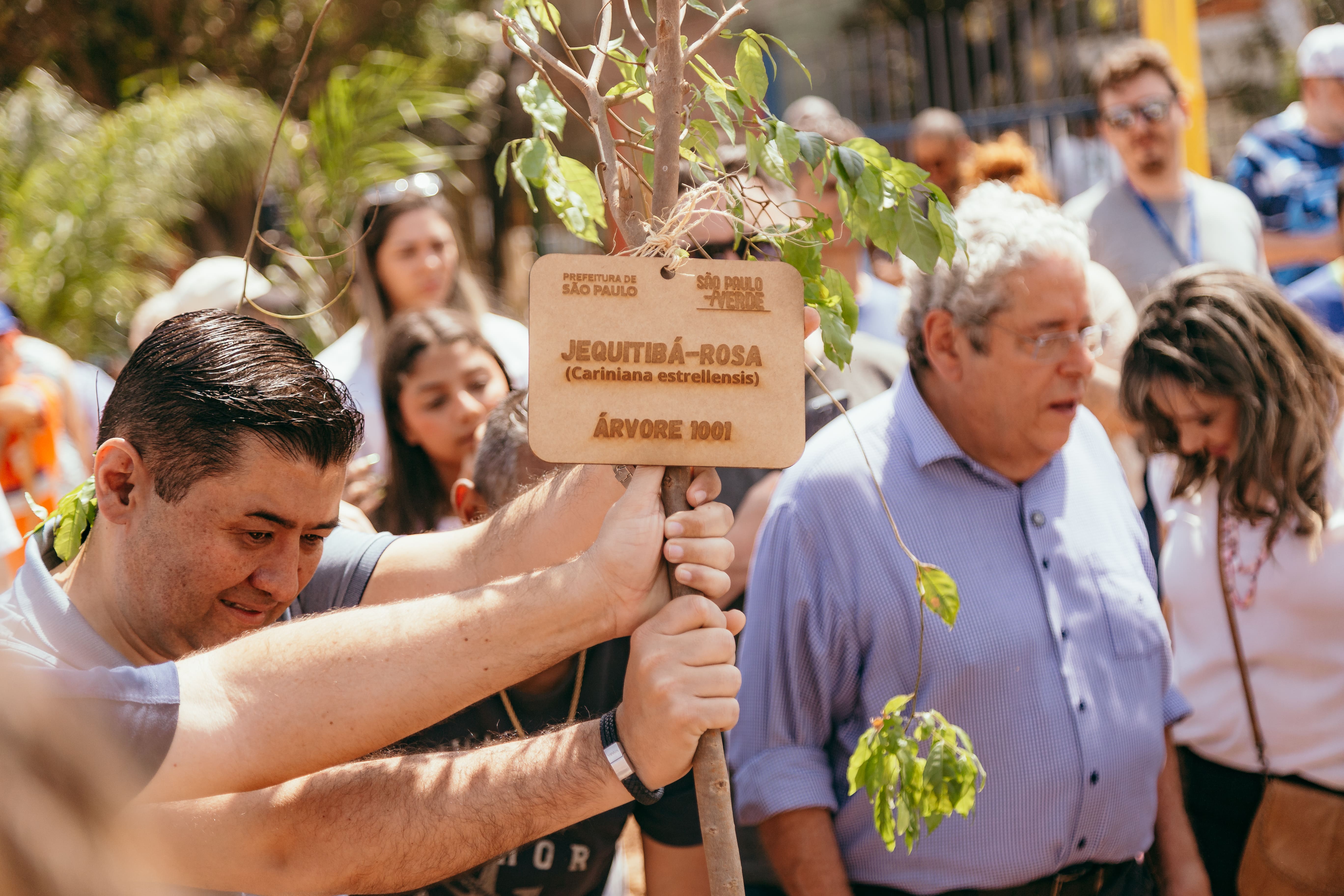 Homem segura uma muda de árvore com uma placa presa ao tronco que identifica a espécie como Jequitibá-rosa (Cariniana estrellensis), árvore número 1001. Ao fundo, várias pessoas acompanham a atividade de plantio em área externa.