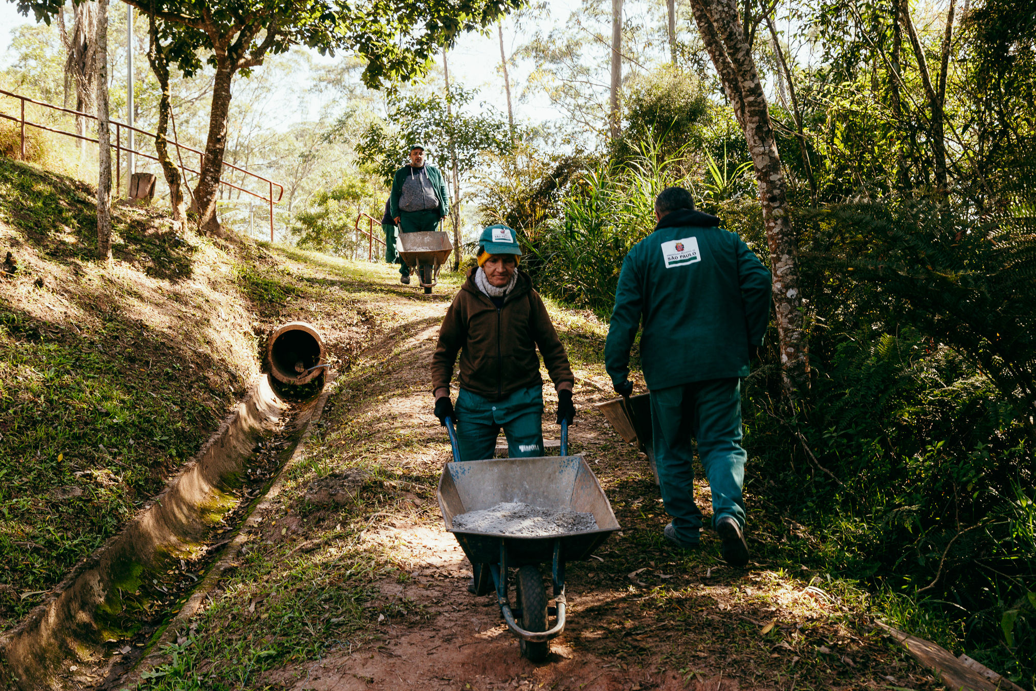 Três trabalhadores vestindo uniformes verdes com o brasão do Governo do Estado de São Paulo caminham por uma trilha em área arborizada. Dois carregam carrinhos de mão com entulho e um terceiro os acompanha. Ao lado da trilha, há uma vala de drenagem e vegetação densa ao redor.
