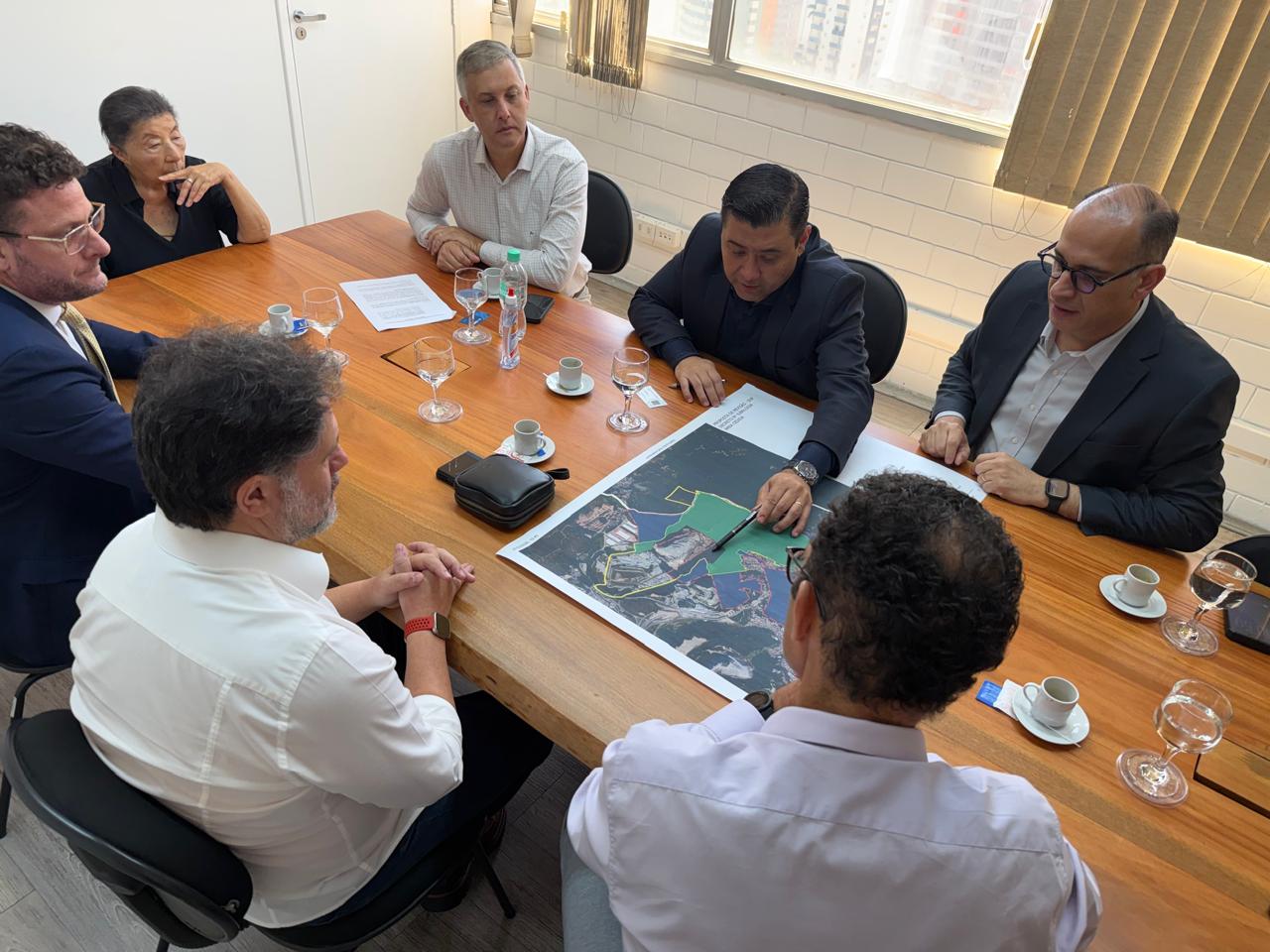 Sete pessoas em uma sala de reunião analisam um mapa sobre a mesa. Um homem aponta para o mapa enquanto os demais observam, com copos, xícaras e documentos ao redor. Ambiente claro e formal.
