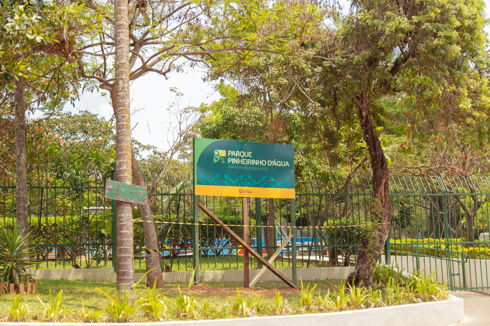 Placa verde e amarela do Parque Pinheirinho D’Água, fixada em frente a uma grade de ferro, cercada por árvores e vegetação. Ao lado, um tronco de árvore traz preso o número “2000”.