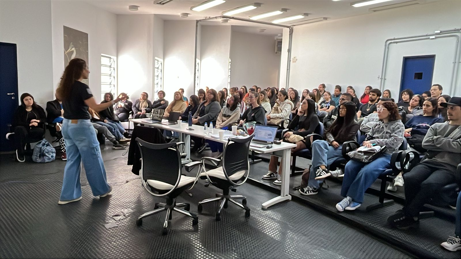 Palestrante em pé diante de um grupo diverso de pessoas atentas em uma sala de aula ou auditório.