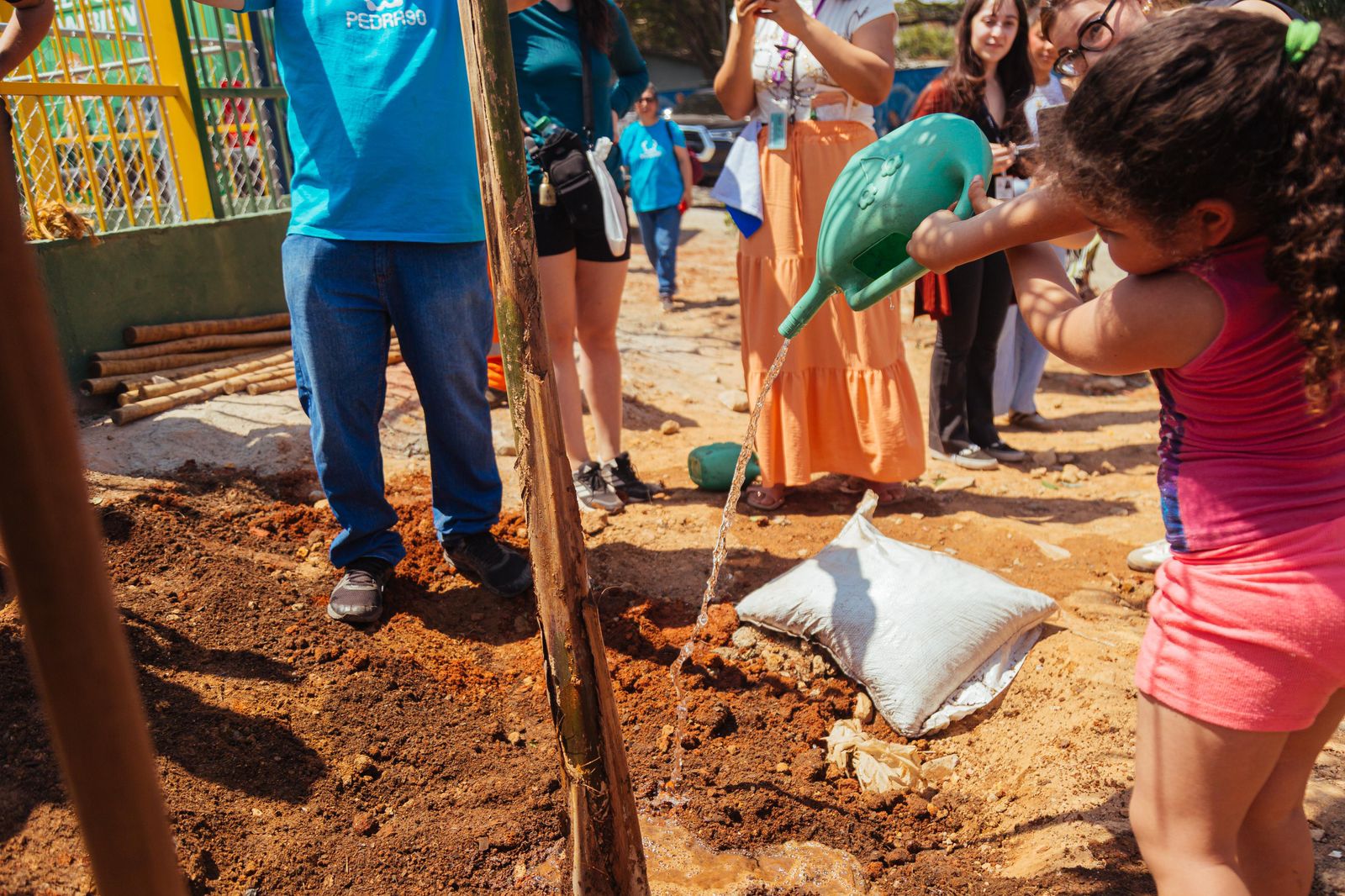 Crianças e adultos participam juntos de um mutirão de plantio em área aberta, em frente a um muro grafitado colorido, com sacos de terra e mudas espalhados pelo chão.