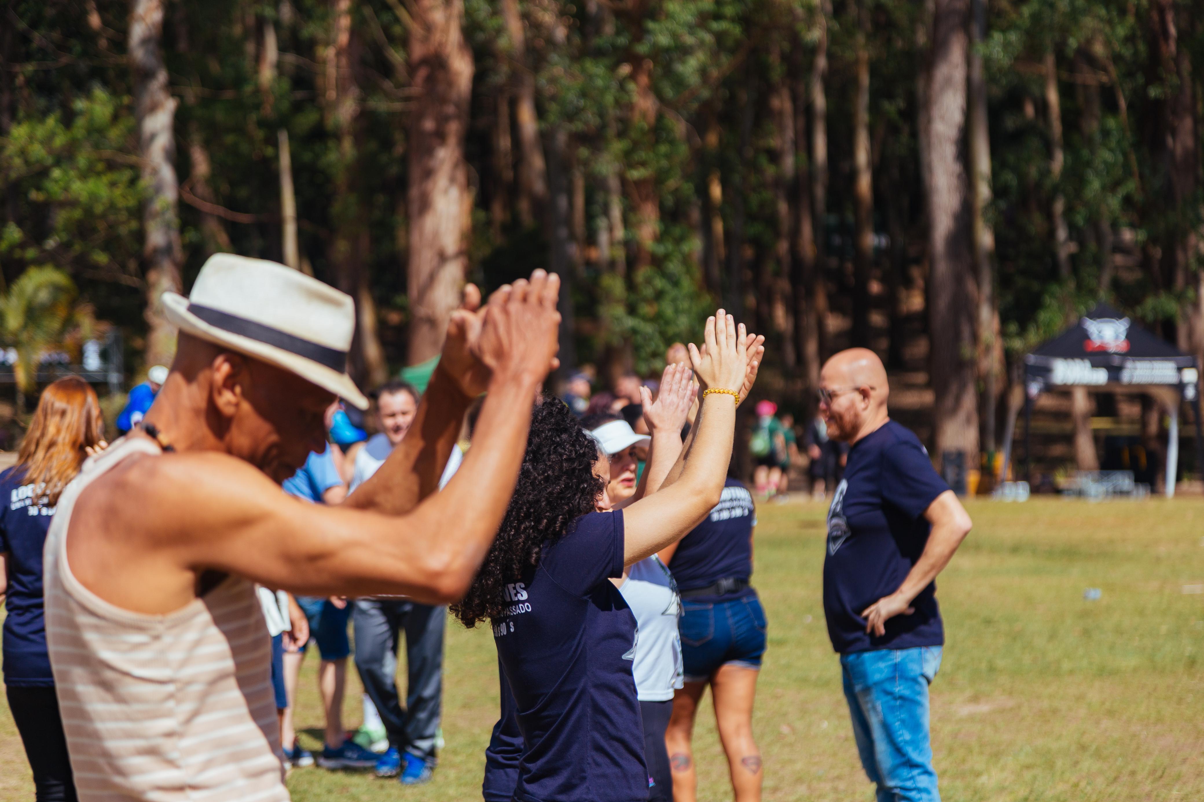 A foto mostra um grupo de pessoas aplaudindo em um evento ao ar livre ensolarado, com uma área gramada e uma floresta densa ao fundo; um homem de pele morena com chapéu panamá branco e regata listrada bege/creme está à esquerda, enquanto uma mulher de cabelo cacheado e camiseta azul marinho com a inscrição "ES PASSADO" está ao lado dele, e um homem careca de óculos e camiseta azul marinho escura observa à direita, com outras pessoas vestindo roupas azuis ao redor, e uma tenda preta com um logotipo branco ao fundo, onde predominam o verde da grama e da vegetação, o azul em várias tonalidades nas roupas, o bege/creme e branco nos acessórios, e o marrom dos troncos das árvores, tudo sob a luz do sol que cria sombras nítidas.