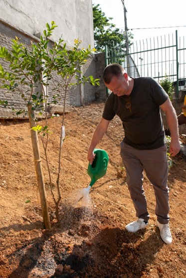 A imagem mostra um homem regando uma muda de árvore em um terreno de terra avermelhada. Ele está inclinado para a frente, segurando um regador verde-claro, de onde sai um jato de água transparente que cai diretamente na base da planta. O homem veste uma camiseta cinza-escura, calça bege e tênis brancos. Ao fundo, aparece um muro cinza e uma cerca metálica verde, além de algumas árvores e vegetação mais distante. A luz do sol é forte, criando sombras definidas e destacando os tons quentes da terra e o verde das folhas da muda.