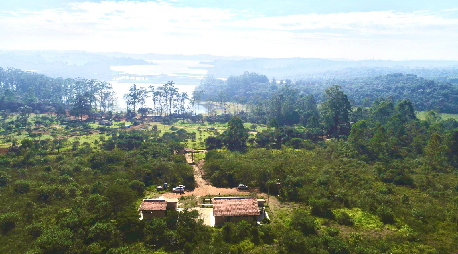Imagem aérea em plano aberto de uma extensa paisagem natural. Em primeiro plano, na parte inferior da imagem, aparecem duas construções térreas com telhados de cerâmica em tom marrom-avermelhado, cercadas por vegetação baixa e arbustos verdes. Um caminho de terra clara, em tons de bege e ocre, corta a área e segue em direção ao fundo da paisagem. Ao redor das edificações, há áreas gramadas em verde claro e escuro, com árvores e pequenos bosques espalhados de forma irregular. No plano médio, a vegetação se torna mais densa, com muitas árvores altas de copas verdes escuras e troncos pouco visíveis. Entre essas árvores, surgem clareiras e áreas abertas em tons de verde mais claro. Ao fundo, é possível ver um grande corpo d’água, com a superfície refletindo a luz do sol em tons esbranquiçados e azulados, parcialmente envolto por uma leve névoa. O horizonte é formado por colinas suaves cobertas por mata, em variações de verde acinzentado devido à distância. O céu ocupa a parte superior da imagem, com tonalidades claras de azul e branco, apresentando nuvens finas e luminosas que reforçam a sensação de amplitude e tranquilidade do cenário.