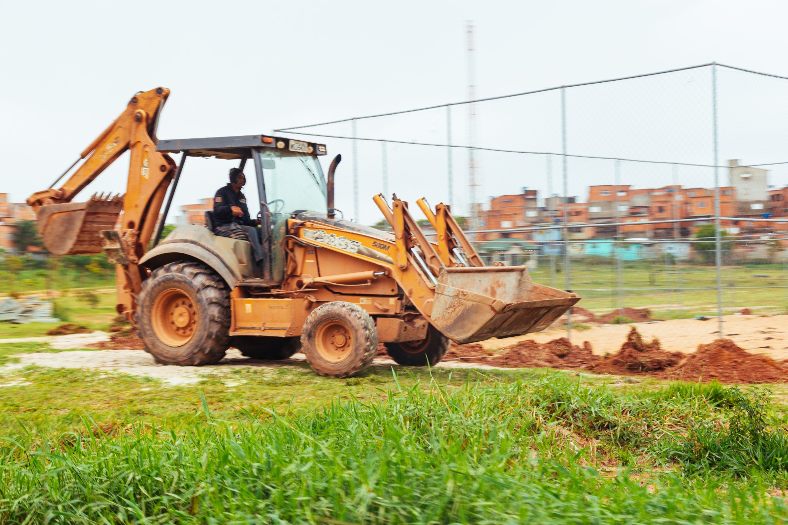Uma retroescavadeira de cor amarela trabalha em uma área aberta, com o operador sentado na cabine. A máquina está sobre o solo de terra avermelhada, com a pá dianteira levantada e a traseira visível. Em primeiro plano há grama verde. Ao fundo, vê-se uma cerca metálica e, mais distante, construções de tijolo em uma área urbana.