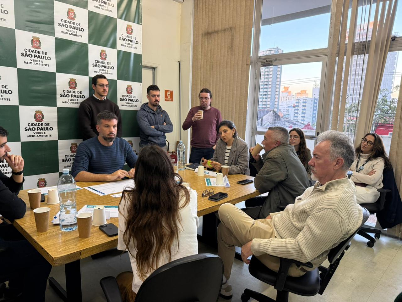 Reunião em sala da Secretaria do Verde e do Meio Ambiente reúne cerca de dez pessoas sentadas e em pé ao redor de uma mesa de madeira. Ao fundo, painel com a identidade visual da Prefeitura de São Paulo. Uma grande janela mostra prédios da cidade ao entardecer.