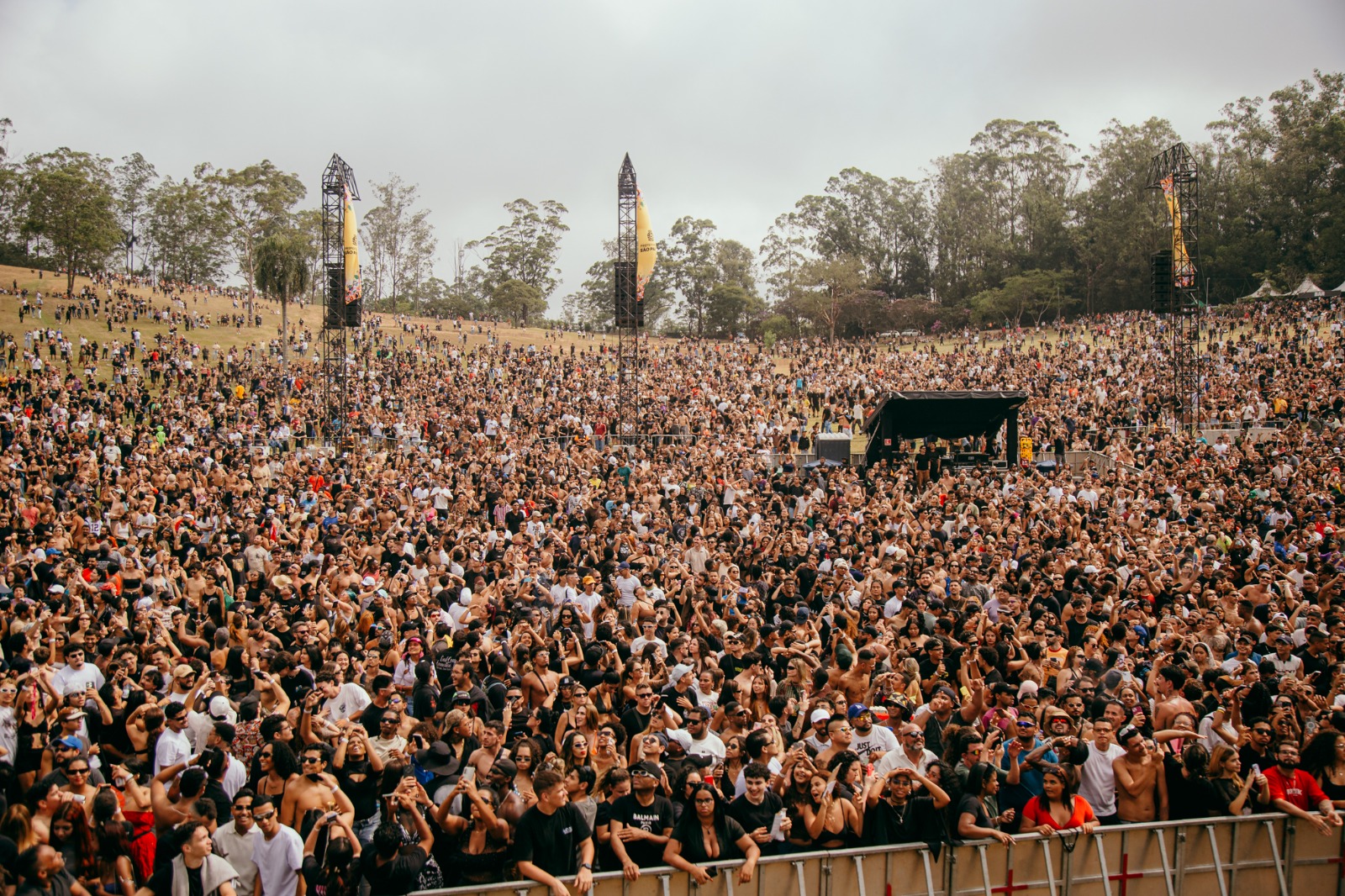Multidão reunida em um grande evento ao ar livre em Parque do Carmo. Milhares de pessoas ocupam a área em frente ao palco e também os gramados ao redor. Ao fundo, há muitas árvores altas e estruturas de som e iluminação. O público aparece animado, com pessoas dançando, levantando os braços e assistindo aos shows durante o dia nublado.