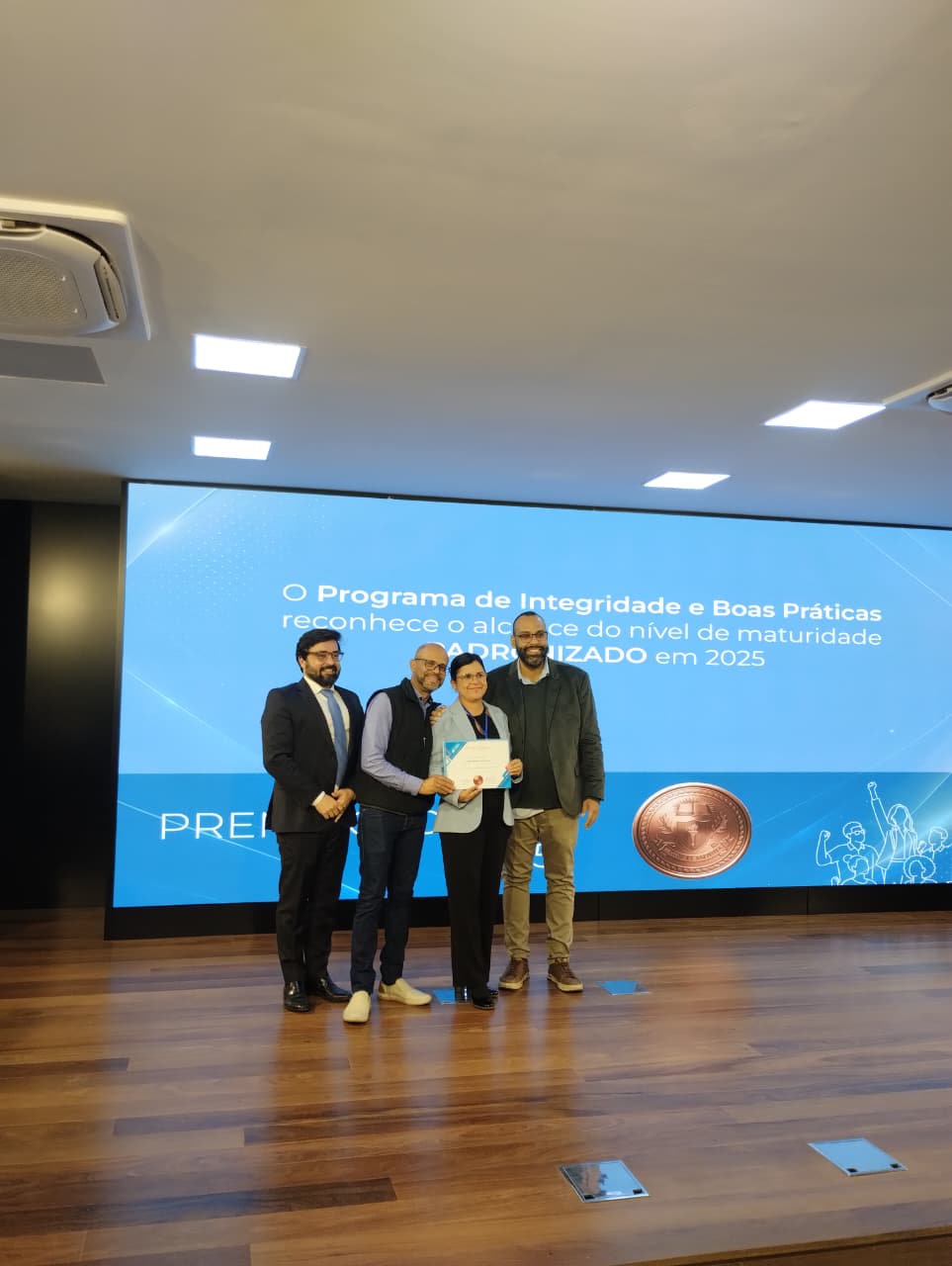 Grupo de pessoas posa em um palco durante uma cerimônia de reconhecimento institucional. Ao fundo, um telão exibe informações sobre o Programa de Integridade e Boas Práticas, enquanto uma das participantes segura um certificado recebido durante o evento.
