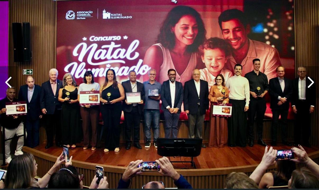 A imagem mostra um grupo de 14 pessoas (homens e mulheres) posicionados lado a lado em um palco de madeira, durante uma cerimônia de premiação. Ao fundo, há um grande painel digital vermelho e brilhante. Na parte inferior da foto, em primeiro plano, aparecem mãos da plateia segurando celulares para registrar o momento.