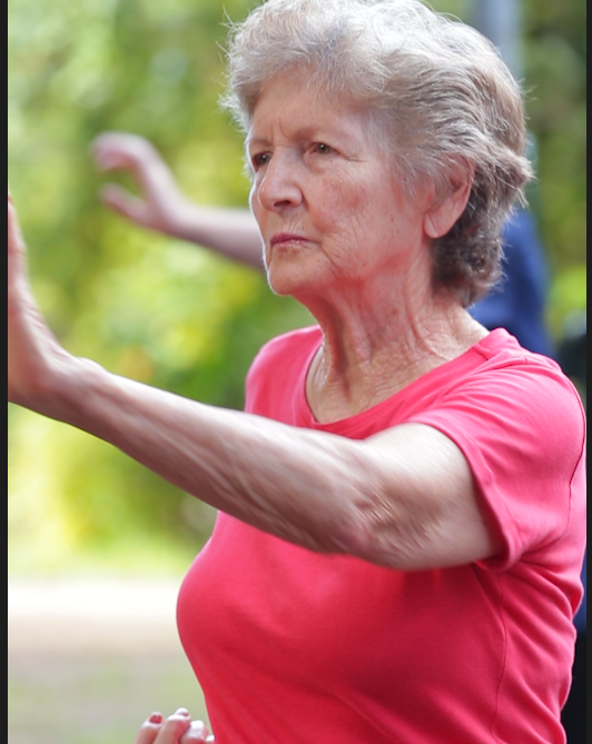 A imagem mostra uma mulher idosa ao ar livre, em um ambiente que parece ser um parque ou área verde, com o fundo desfocado em tons de verde. Ela veste uma camiseta rosa de manga curta e está com os braços erguidos e estendidos à frente, como se estivesse realizando um exercício de alongamento, prática corporal ou movimento de concentração, semelhante a atividades como tai chi, yoga ou ginástica leve.  Seu rosto apresenta uma expressão séria e focada, indicando atenção ao movimento que executa. Os cabelos são curtos, grisalhos, e a postura é ereta, transmitindo equilíbrio e controle corporal. A cena sugere uma atividade física voltada ao bem-estar, à saúde e à manutenção da mobilidade.