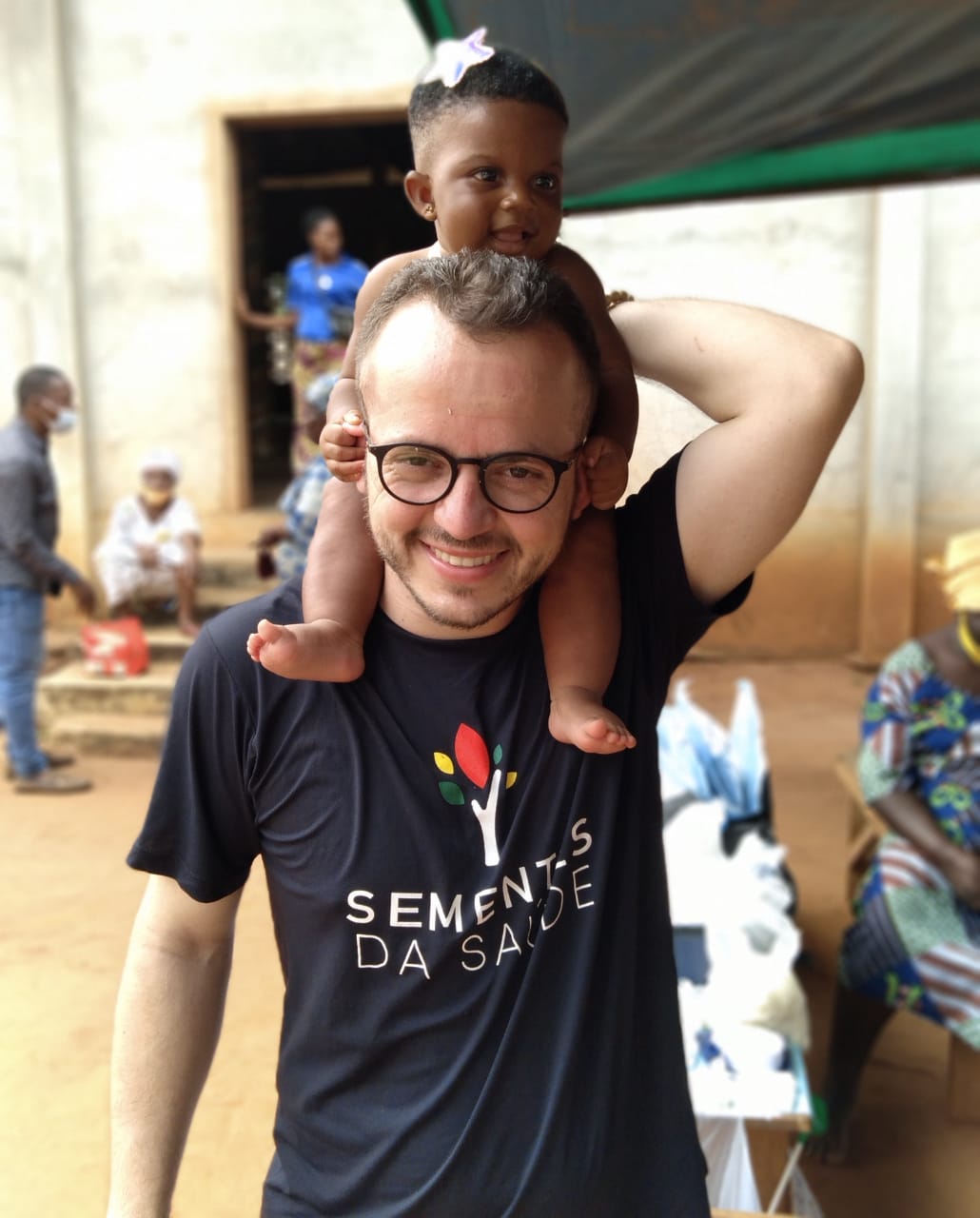 A imagem mostra um homem sorridente usando óculos e uma camiseta preta com o logotipo e a frase “Sementes da Saúde”. Ele carrega uma criança pequena nos ombros, que também sorri alegremente. A criança está descalça, com um laço branco no cabelo e parece divertida com o momento. Ao fundo, há outras pessoas em um ambiente simples, com chão de terra e paredes de concreto, sugerindo uma atividade comunitária ou ação social. A cena transmite afeto, solidariedade e alegria compartilhada.