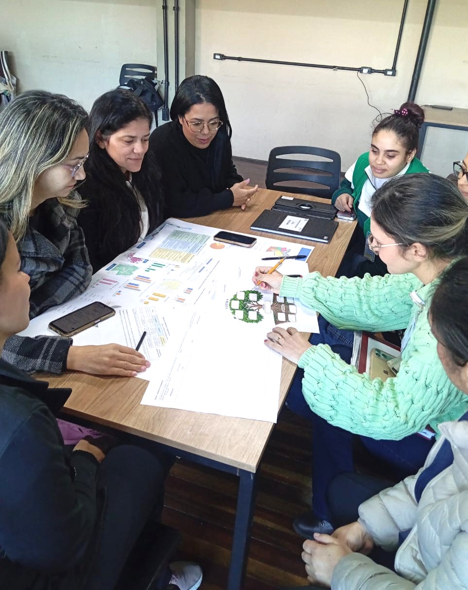A imagem mostra um grupo de pessoas reunidas em torno de uma mesa durante uma atividade colaborativa, aparentemente em um ambiente educacional, comunitário ou de planejamento participativo.  Os participantes estão sentados ao redor da mesa analisando materiais impressos, gráficos, tabelas e esquemas visuais distribuídos sobre uma grande folha de papel. Uma das pessoas desenha ou faz anotações em um diagrama ilustrado, enquanto os demais observam e discutem o conteúdo. Também é possível ver celulares, cadernos e um computador portátil fechados sobre a mesa, indicando um momento de trabalho em grupo ou oficina.