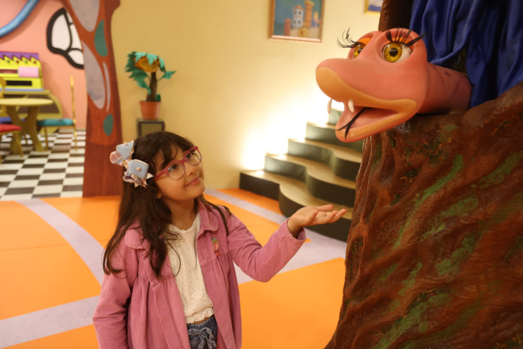 Uma foto de plano médio mostra uma menina com longos cabelos castanhos e óculos de aro rosa, vestindo uma jaqueta de veludo cotelê rosa sobre uma blusa branca. Ela está parada em um ambiente interno colorido que se assemelha a um cenário de programa infantil, com piso de ladrilhos xadrez branco e preto e paredes em tons de laranja, amarelo e verde.  A menina está olhando para cima, para a direita, e estende a mão esquerda aberta em direção a uma marionete de cobra de cor rosa-salmão, que emerge de um tronco de árvore cenográfico texturizado em marrom e verde, no canto superior direito. A marionete tem grandes olhos maquiados com cílios longos e boca aberta mostrando um dente proeminente.  Ao fundo, há uma escada e uma área de estar com móveis coloridos.
