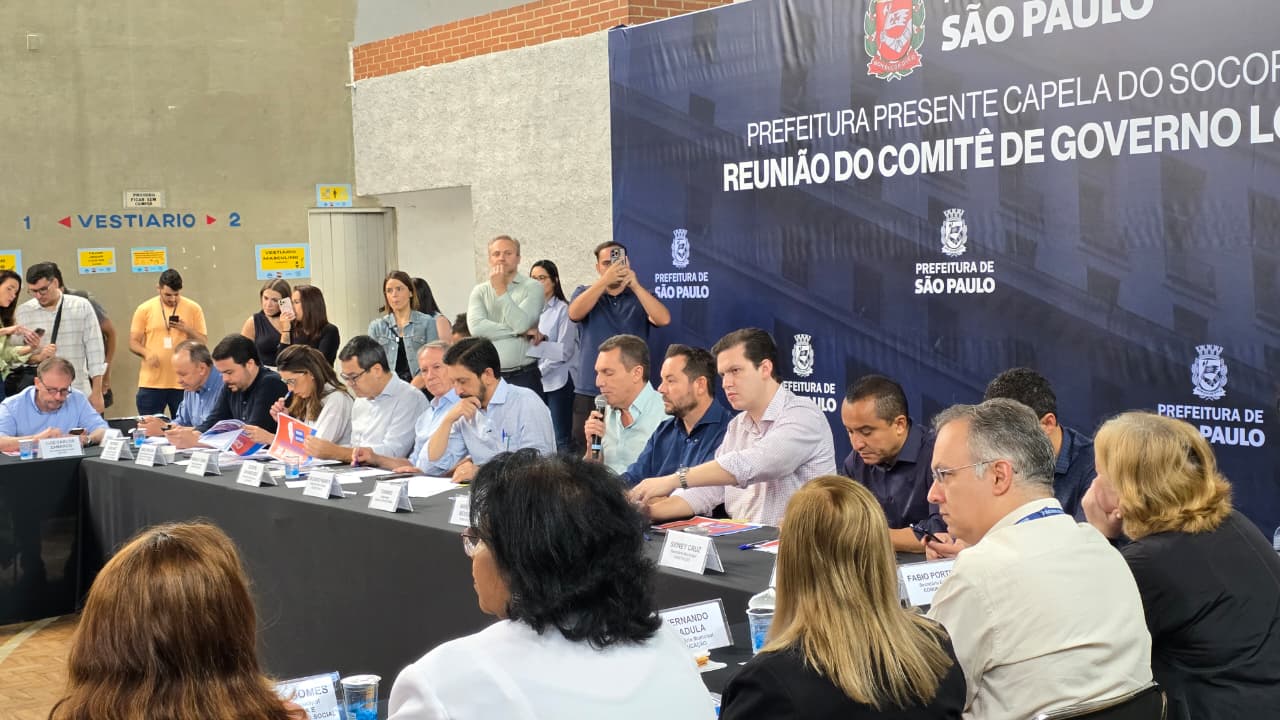 Reunião ampla em um ginásio, com dezenas de representantes sentados ao redor de mesas em formato de “U”. No fundo, um grande painel azul da Prefeitura de São Paulo sinaliza a realização da “Reunião do Comitê de Governo Local”. O espaço está cheio, com participantes acompanhando atentos e outras pessoas de pé ao redor registrando e observando o encontro.