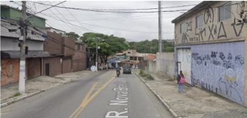  Foto de uma rua em São Paulo, Brasil, chamada Rua Nova Brasil. A rua é asfaltada e tem uma faixa amarela no centro. Há construções de um e dois andares em ambos os lados da rua, algumas com fachadas simples e outras com grafites. Uma mulher com uma mochila rosa está caminhando na calçada à direita. Um carro preto e uma motocicleta estão dirigindo na rua, em direção à câmera. Fios elétricos cruzam o céu nublado. Há árvores e vegetação visíveis entre as construções. A foto parece ter sido tirada durante o dia, sob luz natural difusa.