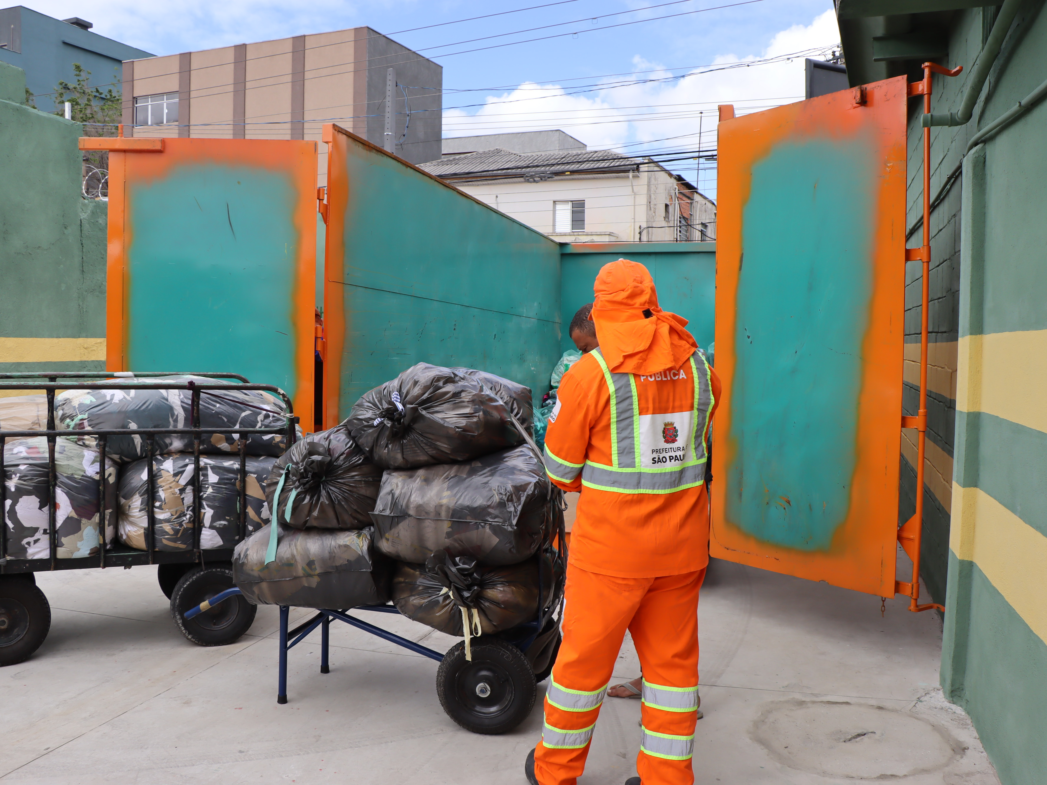 Fotografia em um pátio ou área de serviço, mostrando a coleta de resíduos. Em primeiro plano, um trabalhador da limpeza urbana está de costas, vestindo um macacão e colete de segurança de cor laranja vibrante com faixas refletivas. No colete, é visível o logotipo e a inscrição "PREFEITURA DE SÃO PAULO".  O trabalhador está próximo a duas carroças de mão carregadas com grandes sacos plásticos pretos e sacos camuflados. As sacolas estão cheias, presumivelmente de resíduos têxteis ou outros materiais leves, amontoadas sobre as carroças.  Ao fundo, há um grande portão aberto, com a parte interna pintada em um tom de verde-água e as bordas em laranja queimado. Pelo portão, é possível ver um muro amarelo e preto à direita e construções urbanas e um céu azul com nuvens brancas no horizonte.
