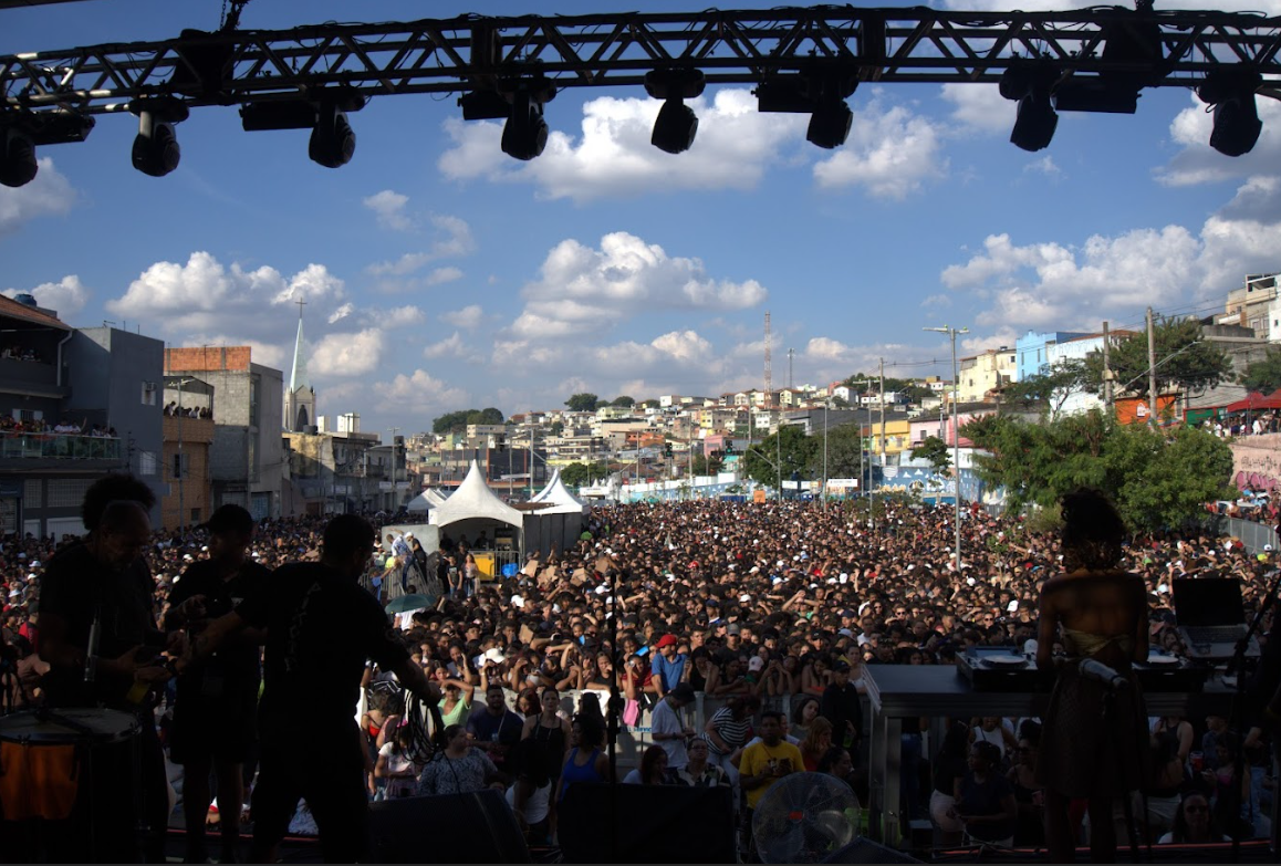 Uma vista ampla de um grande público em um evento ao ar livre, possivelmente um show ou festival, com uma estrutura de palco na parte superior da imagem, de onde a foto parece ter sido tirada. A multidão preenche a maior parte do campo de visão, estendendo-se ao longe. Ao fundo, uma paisagem urbana com muitas casas e edifícios em uma encosta, alguns deles coloridos, sugerindo uma área residencial. O céu é azul com nuvens brancas dispersas, indicando que o evento ocorre durante o dia. Há também algumas tendas brancas visíveis entre a multidão. Em primeiro plano, silhuetas de pessoas no palco, possivelmente artistas ou membros da equipe, são visíveis.