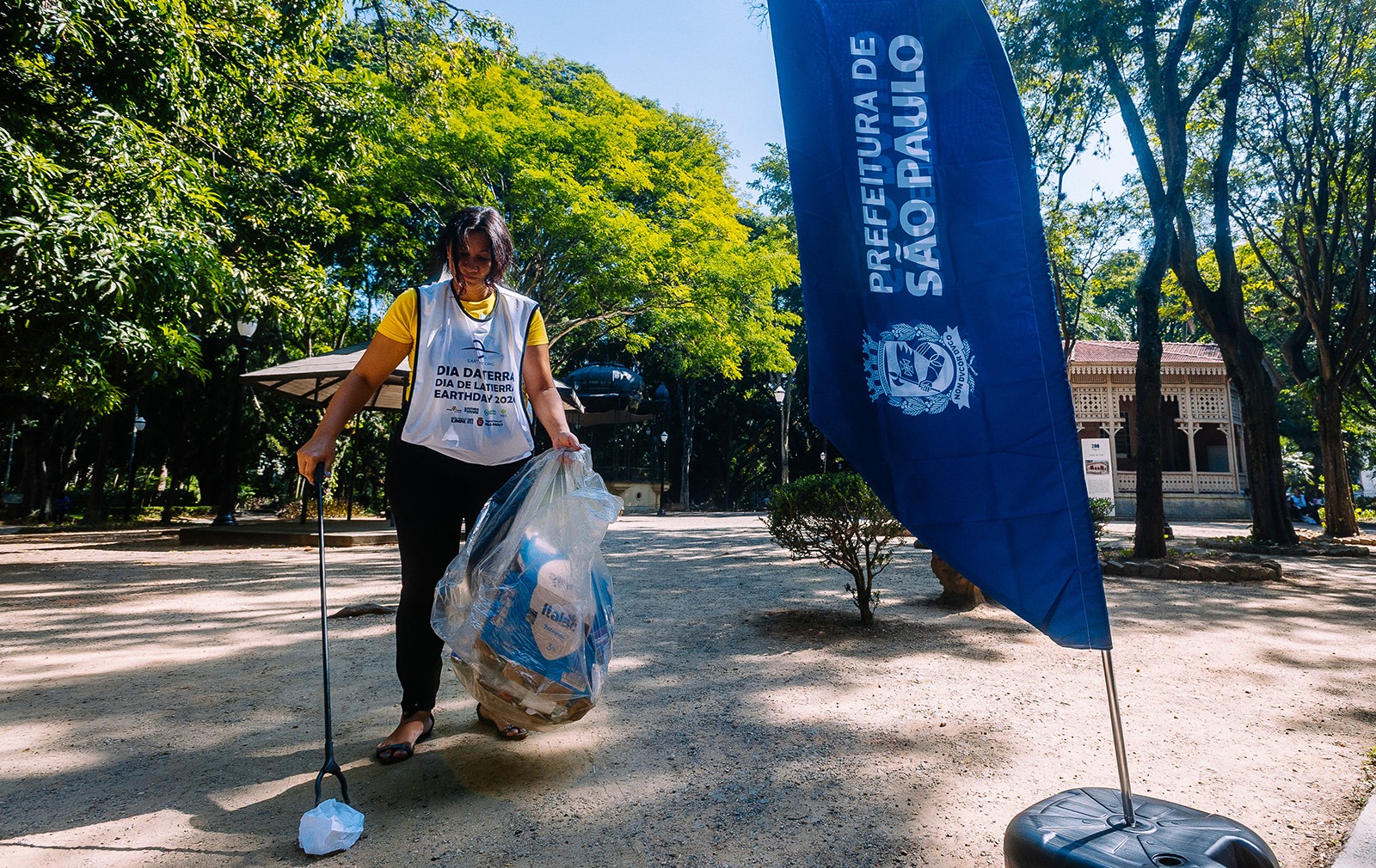 Fotografia colorida em um parque ensolarado. No centro, uma mulher parda de cabelos escuros veste camiseta amarela e um colete branco de voluntária com os dizeres "Dia da Terra / Earth Day 2024". Ela está levemente inclinada, usando um pegador longo para recolher um papel do chão, enquanto segura um saco plástico transparente com recicláveis na outra mão. À direita, em destaque, aparece uma bandeira vertical azul com o logotipo da Prefeitura de São Paulo. Ao fundo, árvores altas com copas verdes e um coreto de madeira completam a cena sob a luz forte do sol, que projeta sombras nítidas no caminho de terra.